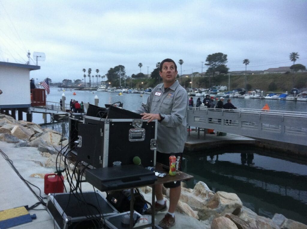 Dave is wearing his name shirt and is standing behind a soundboard with the ocean in the background.